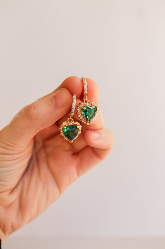 A hand holds two heart-shaped earrings featuring green gemstones, surrounded by gold embellishments and small, sparkling stones, against a plain, light background.