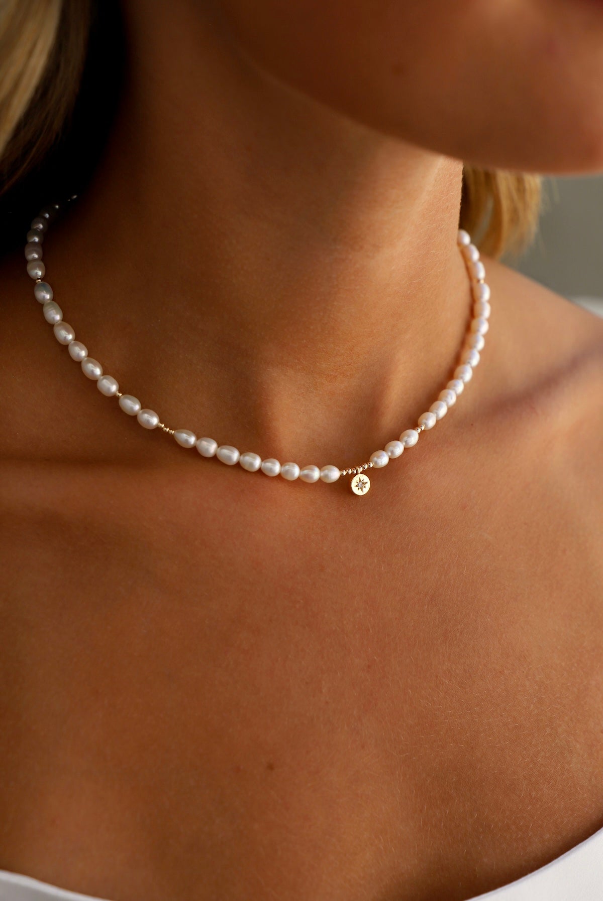 A pearl necklace adorns the neck of a person, featuring a small gold charm. The skin tone is visible, emphasizing elegance against a neutral background.