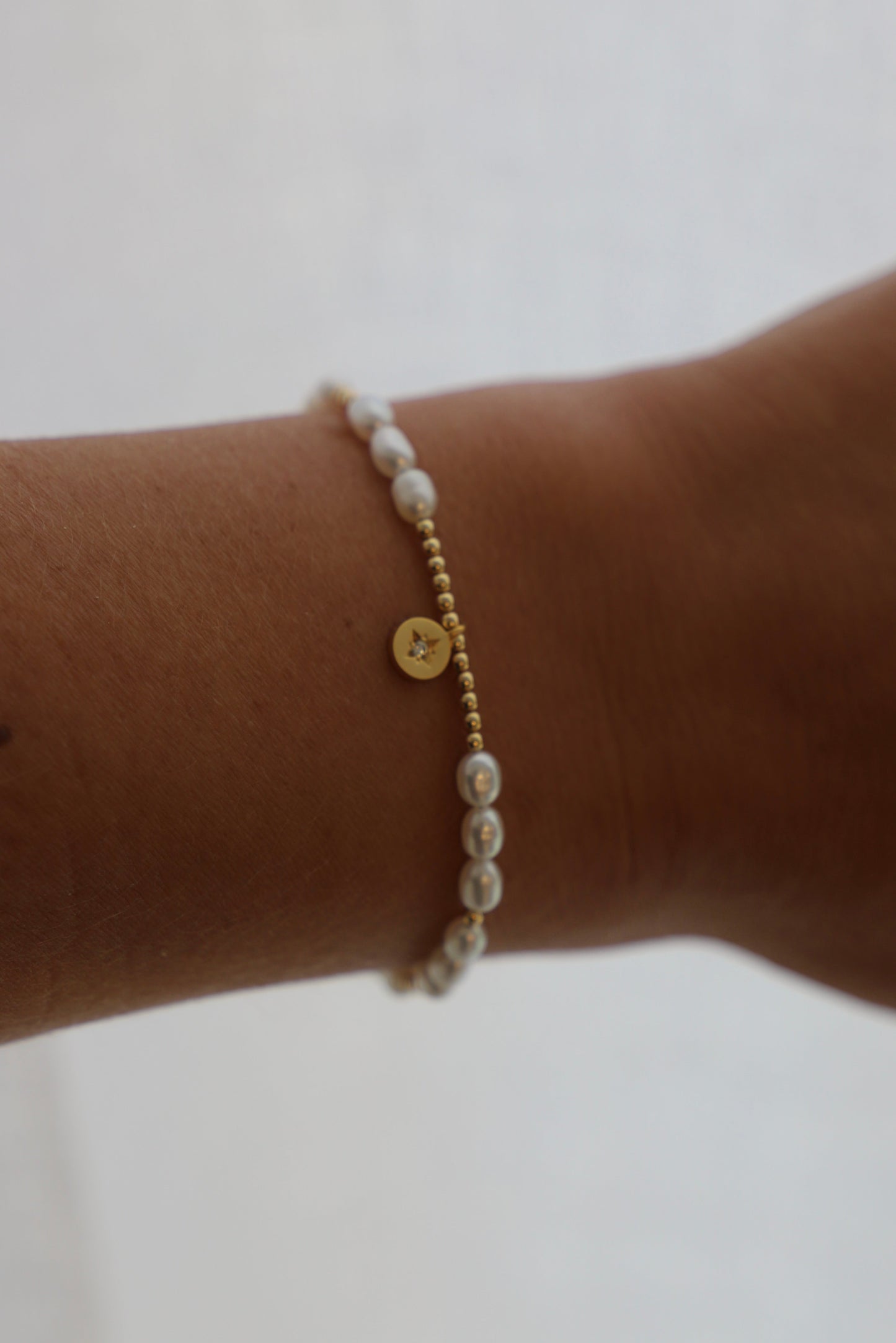 A bracelet is worn on a wrist, featuring alternating small gold beads and white pearls, with a small gold charm at the center, set against a soft, light background.