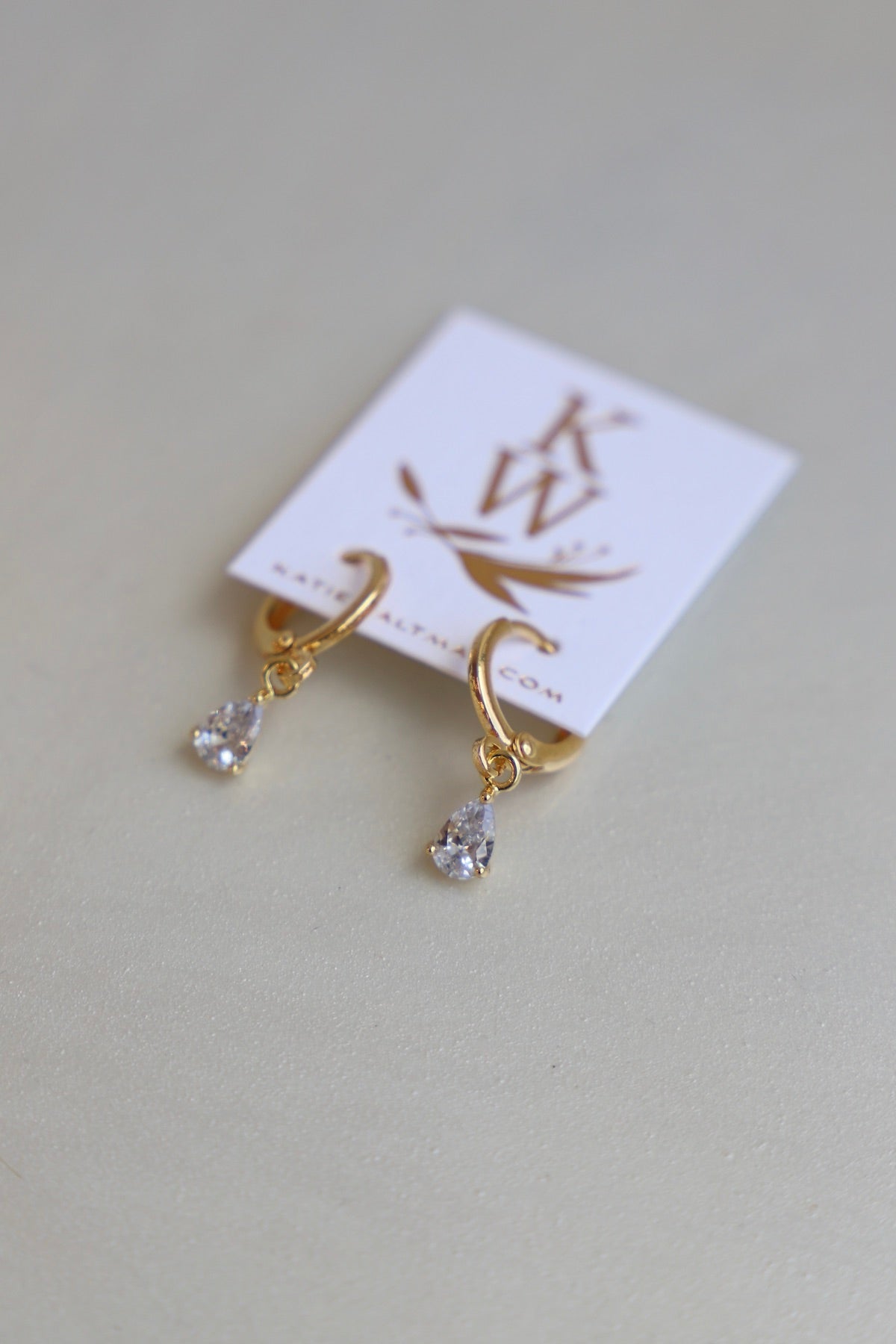 Elegant gold hoop earrings with teardrop-shaped clear crystals hang from a branded card, resting on a smooth, light-colored surface, surrounded by a soft, neutral ambiance.