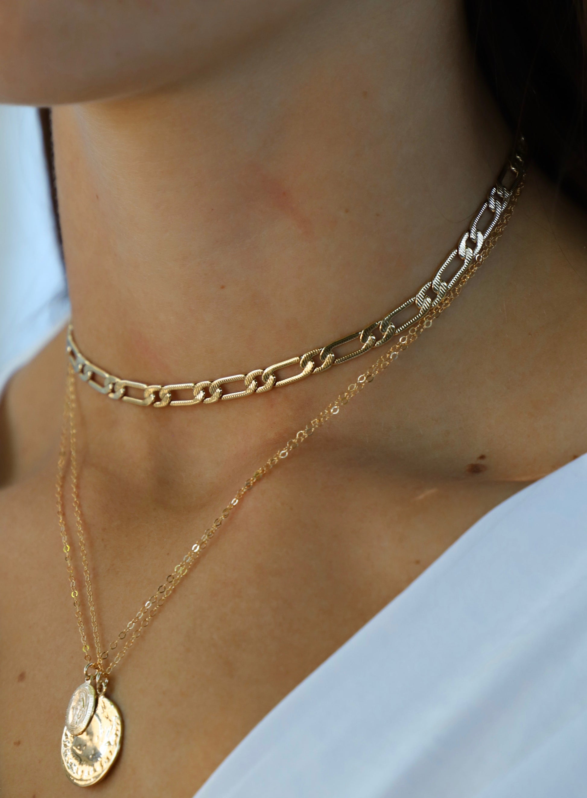 A layered arrangement of gold necklaces adorns a person's neck. The chains include a chunky choker and delicate pendants, set against smooth, fair skin, while partially visible clothing is white.