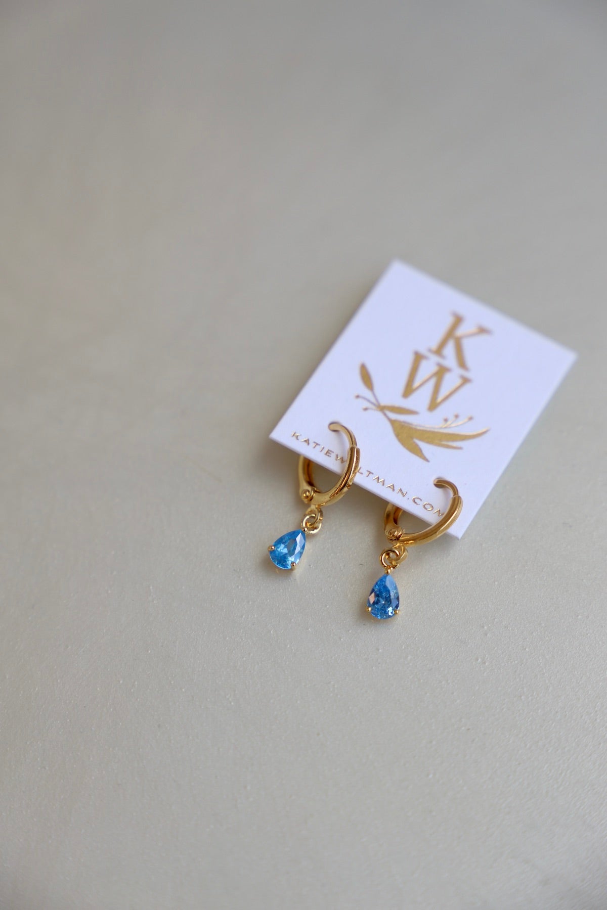 Gold hoop earrings with dangling blue gemstones are displayed on a white card featuring a logo and website URL, set against a light, neutral background.