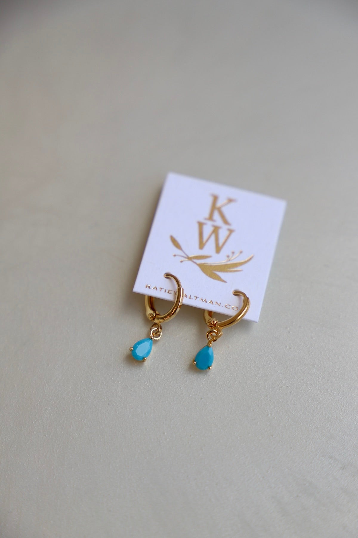 Gold hoop earrings with turquoise teardrop charms rest on a light-colored surface. A small, white card with gold lettering displays the brand name "KW" and the website "katiealtman.com."