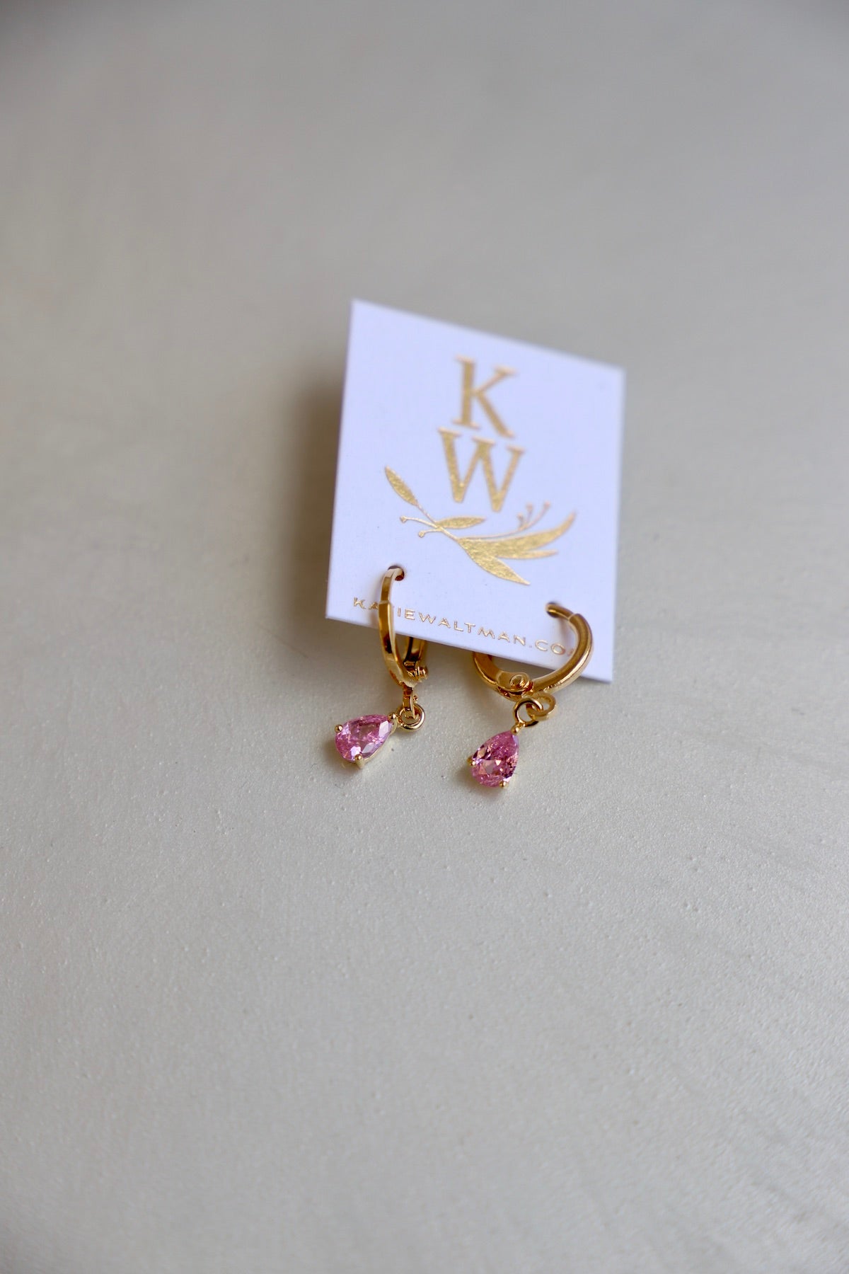 A pair of gold hoop earrings with pink teardrop gemstones hangs from a white card displaying "KW" and "kathiewaltman.com" in gold lettering, set against a light surface.