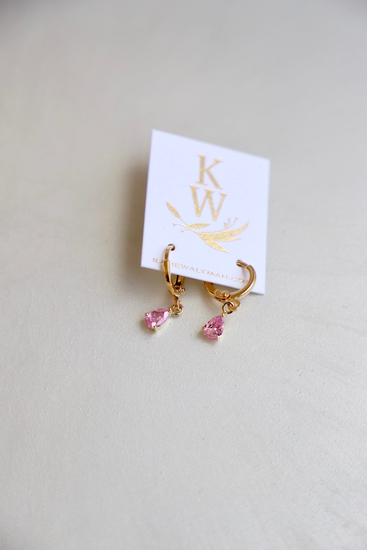 Gold hoop earrings with pink teardrop stones hang from a white cardstock tag featuring the initials â€œKWâ€ in gold, resting on a light-colored surface.