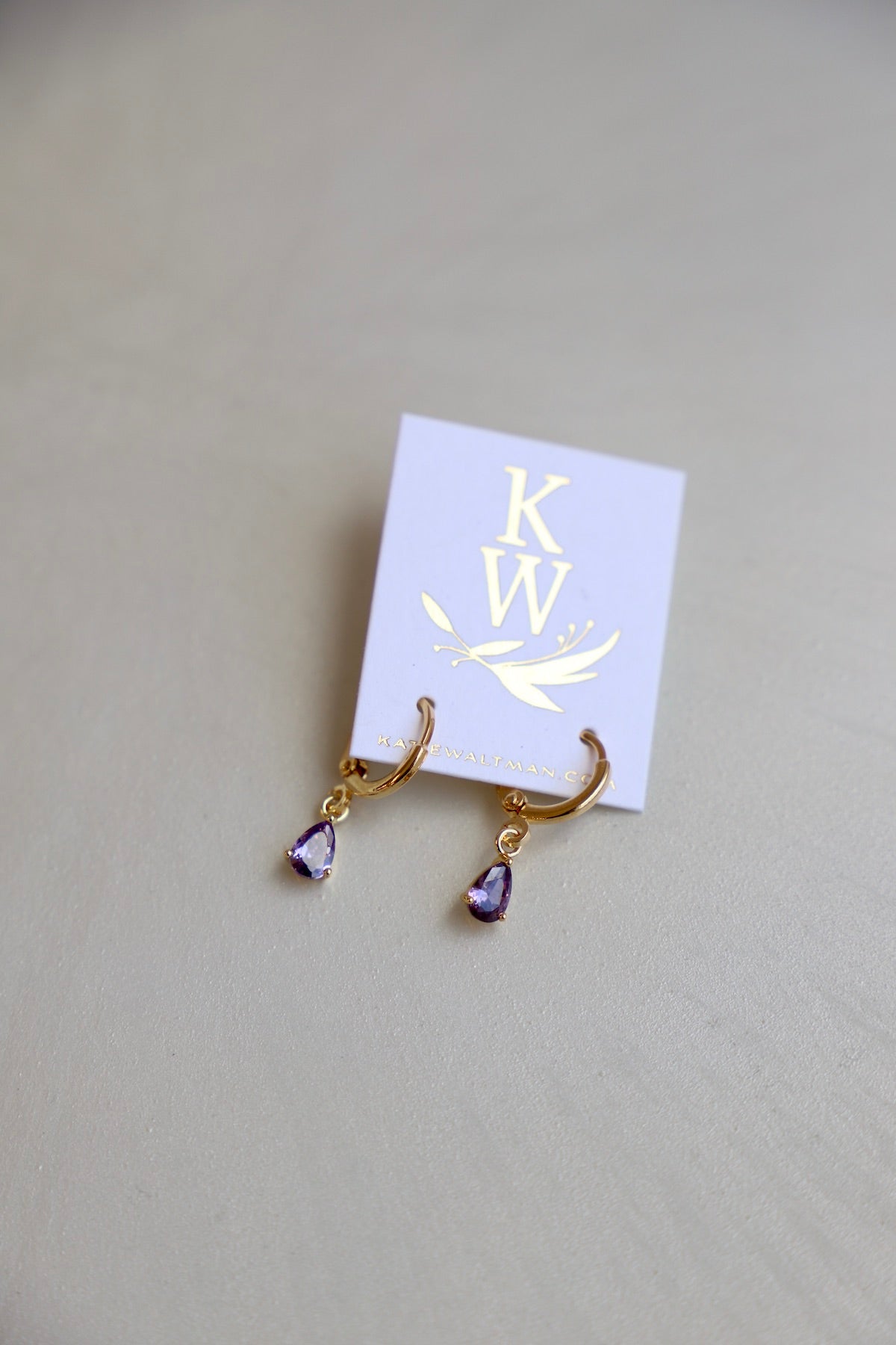 Golden hoop earrings with purple teardrop gemstones hang from a branded card. The card features a gold logo on a light purple background, resting on a smooth beige surface.