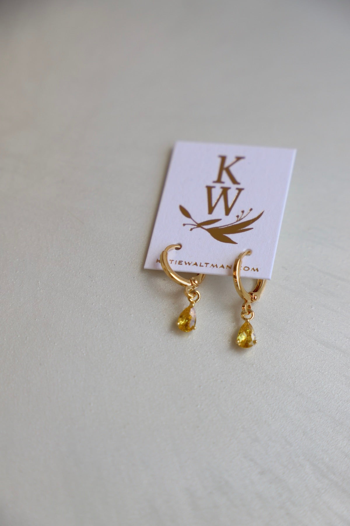 Gold hoop earrings with yellow teardrop gemstones hang from a white card displaying the initials "KW" and a decorative design. The card is placed on a light, textured surface.