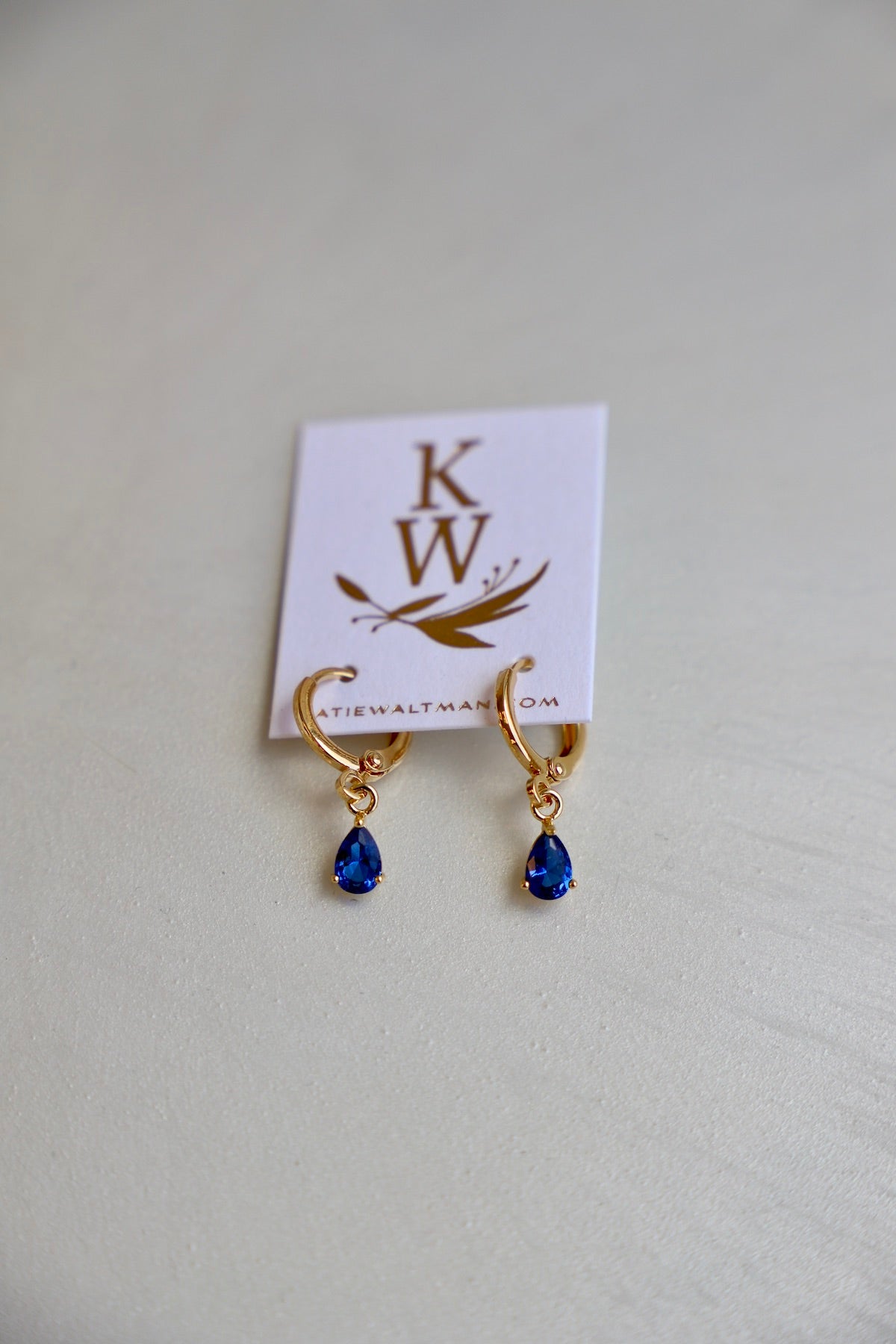 Golden hoop earrings with blue teardrop gems dangle from a white card. The card features the initials "KW" and the website "katiewaltman.com." The surface is softly textured.