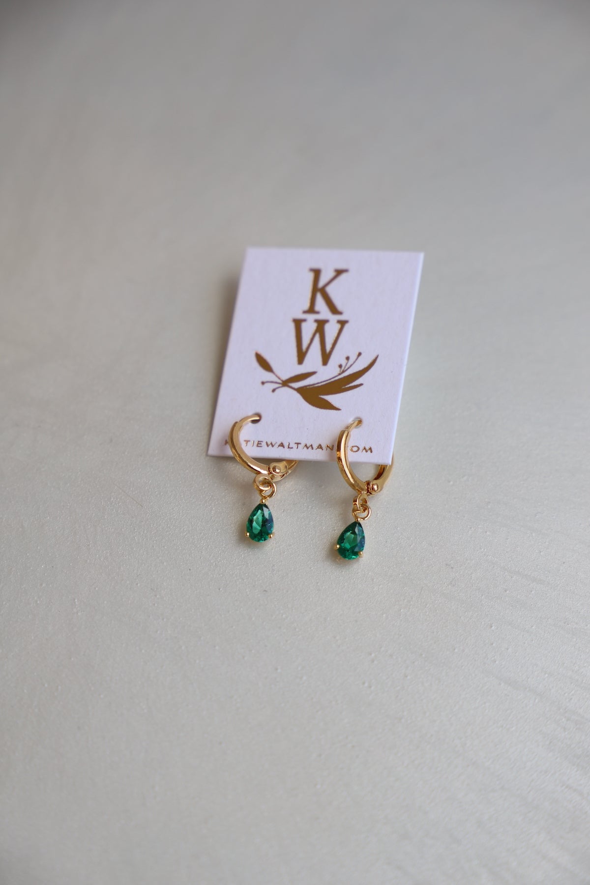 Gold hoop earrings with green teardrop gemstones are displayed on a white card featuring the initials "KW" and the website "katiewaltman.com," placed on a light-colored surface.