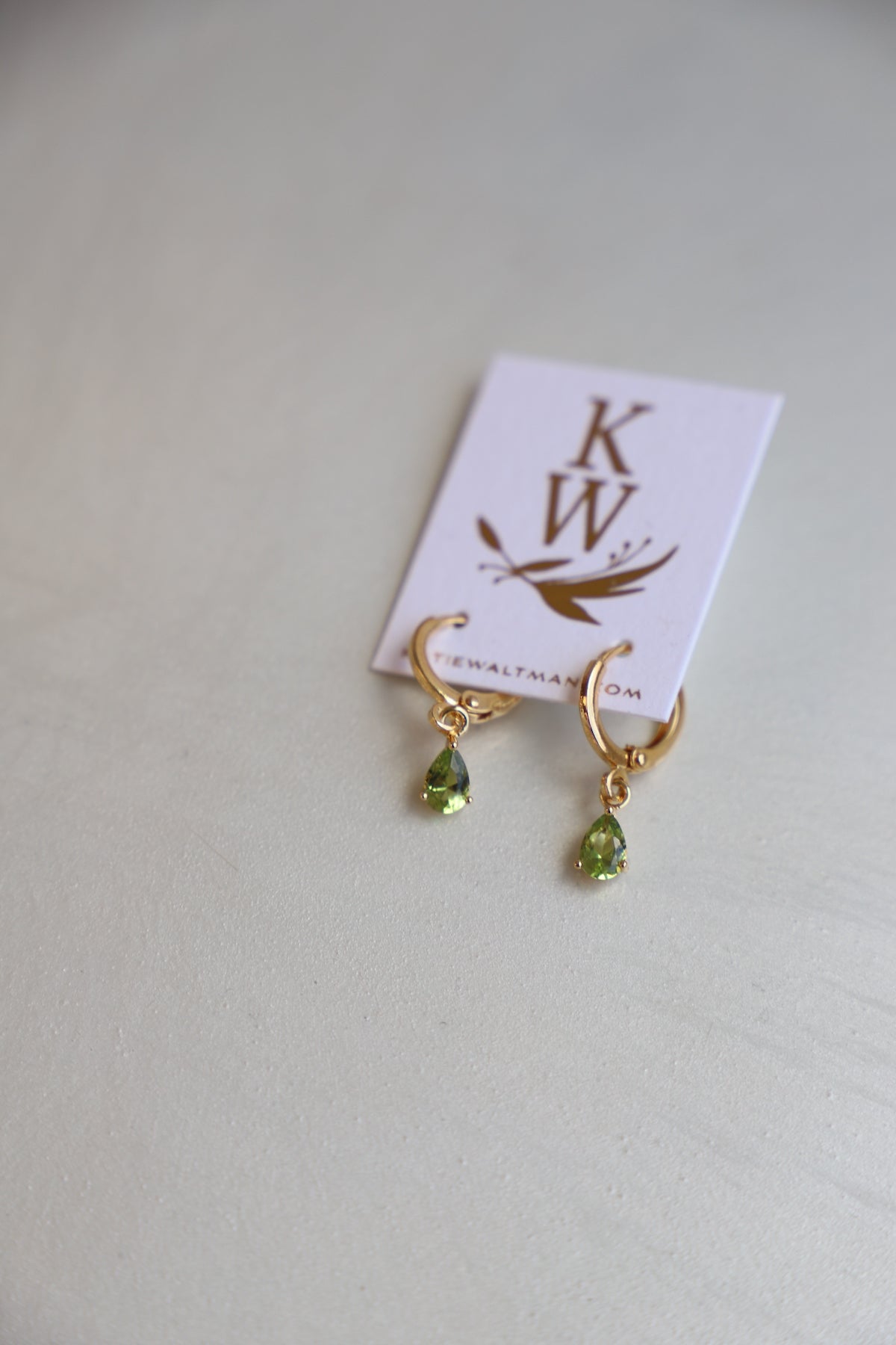 Gold hoop earrings with green teardrop stones hang from a white card displaying a logo. The setting is a light-colored surface, providing a subtle, elegant backdrop.