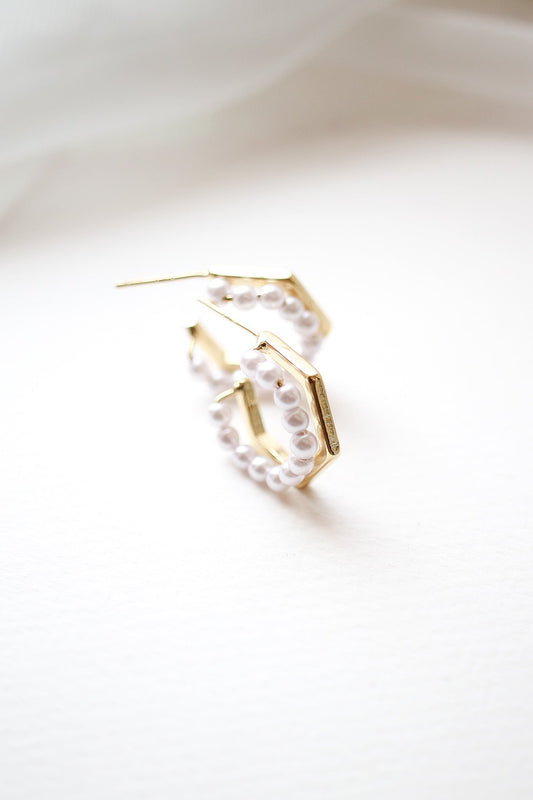 Gold hoop earrings are positioned upright, adorned with small white pearls. They rest on a textured, light-colored surface, highlighting their elegant design against a soft, blurred background.