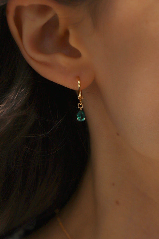 A gold hoop earring with a dangling teal gemstone is worn on the lobe of a woman's ear, framed by long dark hair against a neutral background.