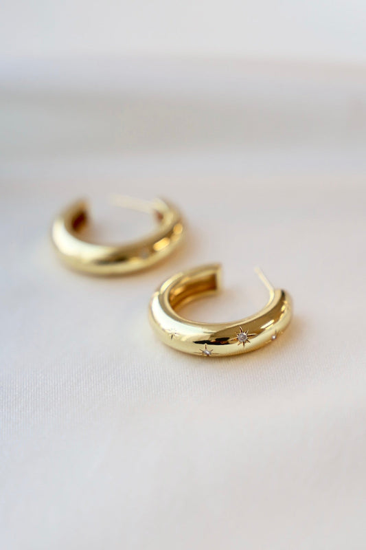 Two gold hoop earrings with star-shaped embellishments rest on a soft, light-colored fabric background, showcasing their polished surface and decorative details in a serene and elegant context.