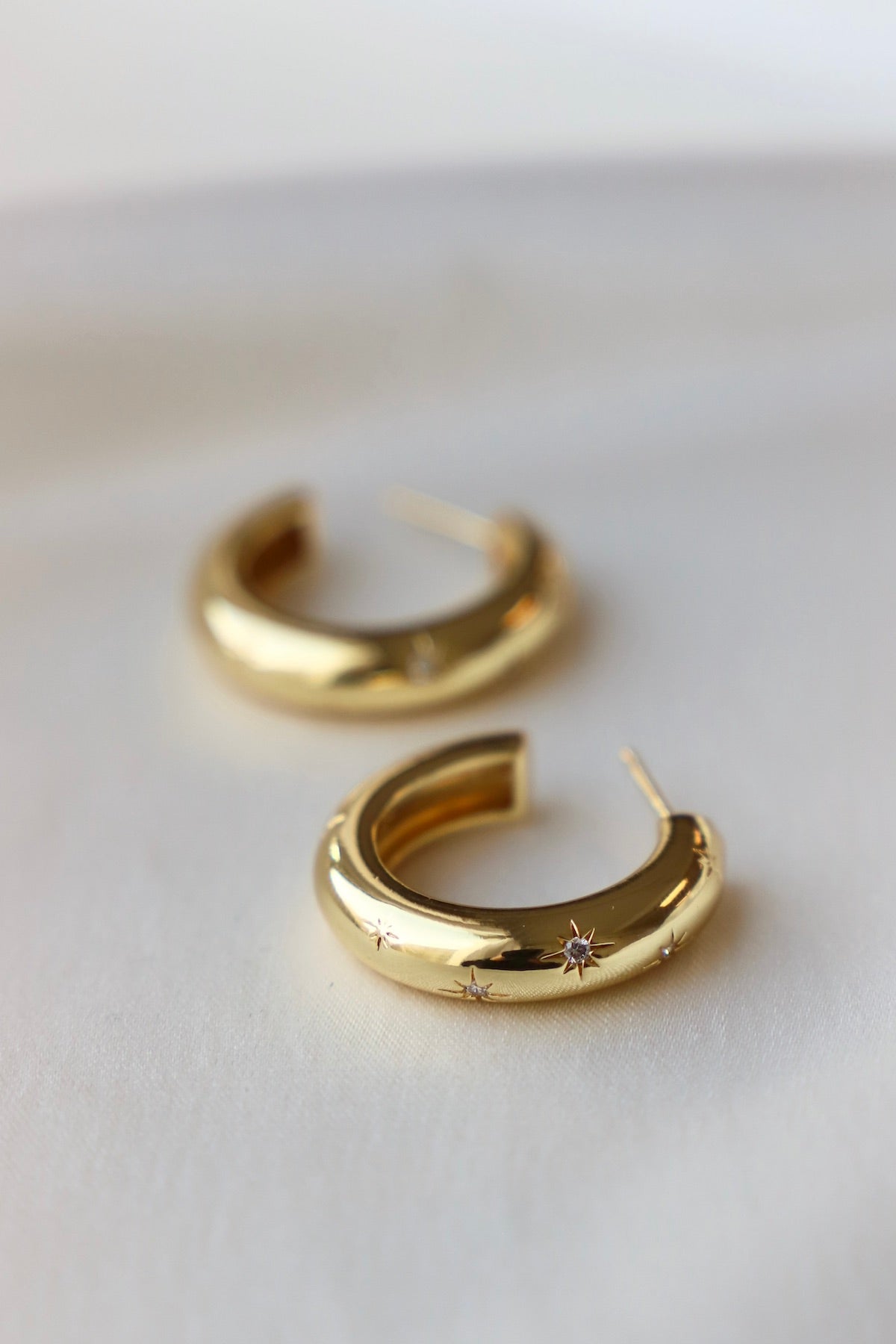 Gold hoop earrings sit on a smooth, light-colored surface, showcasing a polished finish with small star-like embellishments, reflecting soft natural light in a minimalistic setting.