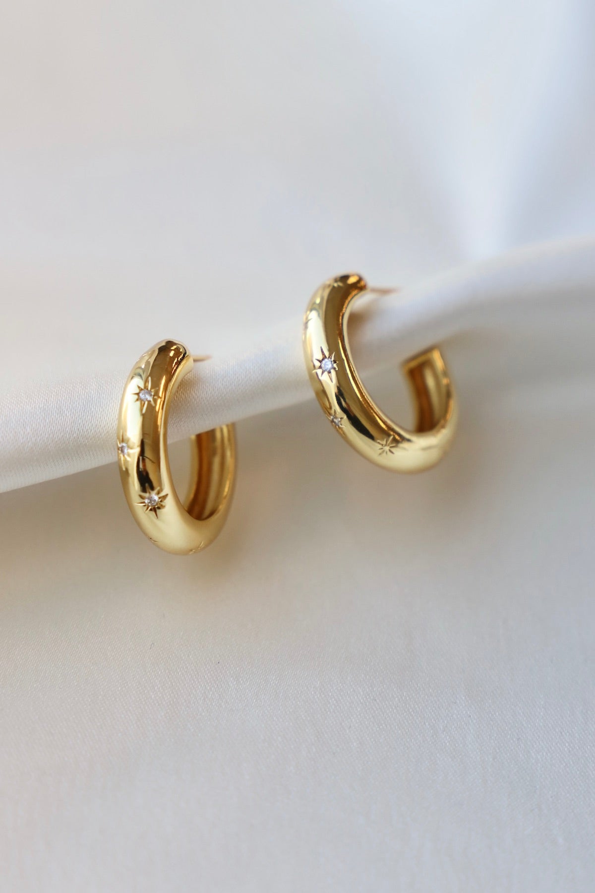 Two gold hoop earrings, adorned with small star-like designs, are resting on a smooth, pale fabric surface, showcasing their shiny finish and elegant curvature.