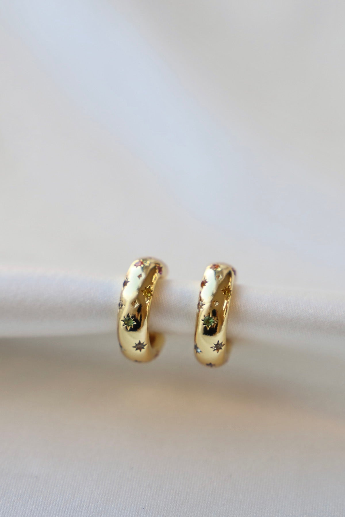 Two gold hoop earrings with decorative star and floral patterns rest on a light-colored fabric surface, highlighting their shine and intricate design in a soft, elegant setting.