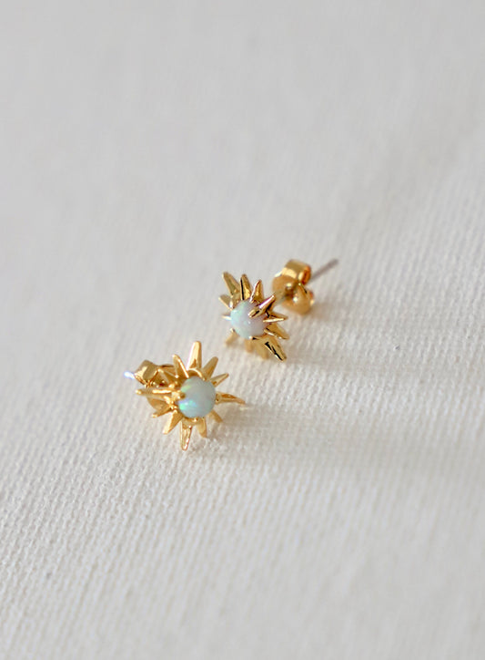 Two golden star-shaped earrings are resting on a light, textured fabric. Each earring features a central opal stone, reflecting subtle colors in a softly lit environment.