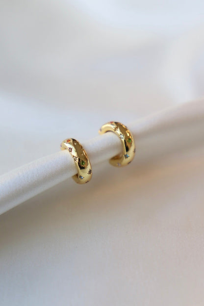 Two gold hoop earrings, adorned with colorful star motifs, rest on a white fabric surface, showcasing their polished surface in a softly lit, minimalistic setting.