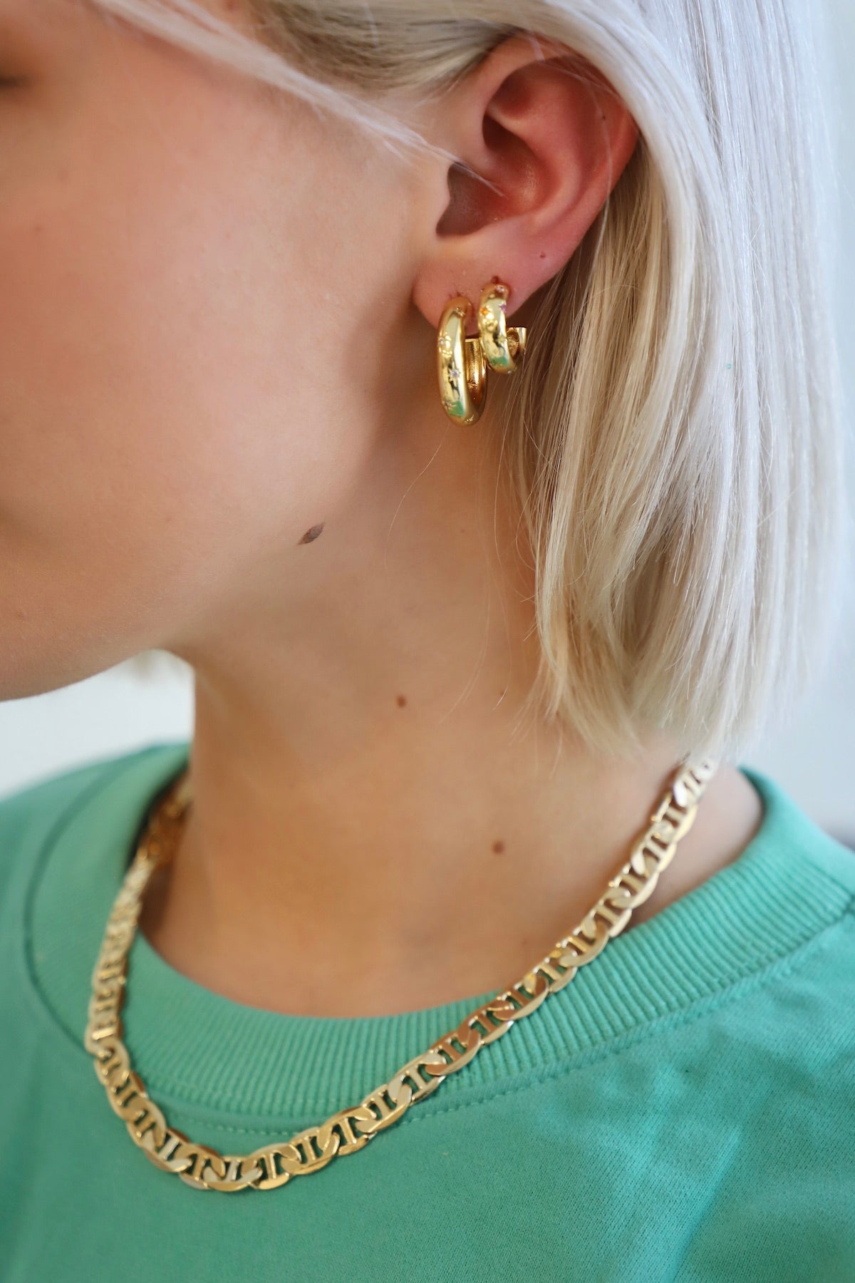 A close-up of a person's ear adorned with bold gold hoop earrings, complemented by a chunky gold chain necklace, set against a soft, light-colored background and wearing a mint green shirt.