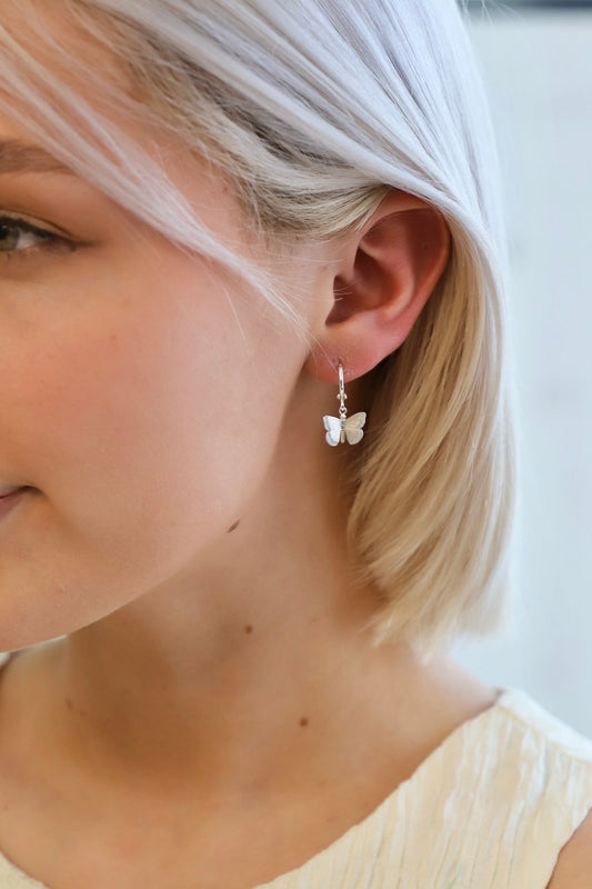 A silver butterfly earring hangs from the ear of a young woman with short, light blonde hair. She is partially smiling, set against a softly lit background.