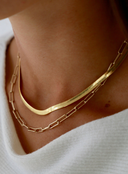 Two gold necklaces are worn around a neck, with a flat, curved chain on top and a linked chain below. The skin tone contrasts with the white, textured garment underneath.