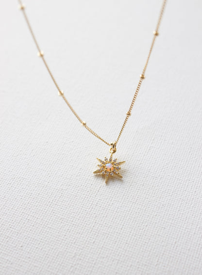 A gold necklace features a star-shaped pendant with an iridescent center, resting against a textured white background, emphasizing its delicate design and shimmer.