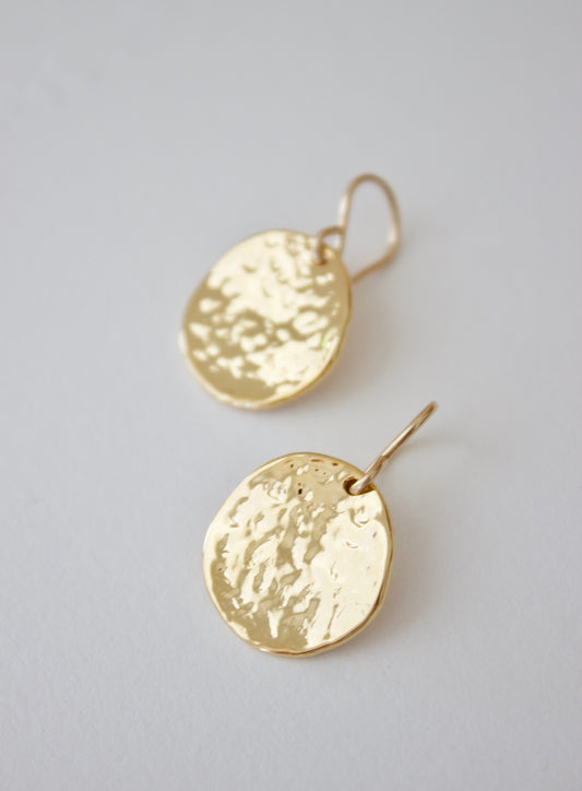 Two golden earrings with a hammered texture hang from thin hooks, resting on a light-colored surface that highlights their reflective surfaces and irregular shapes.