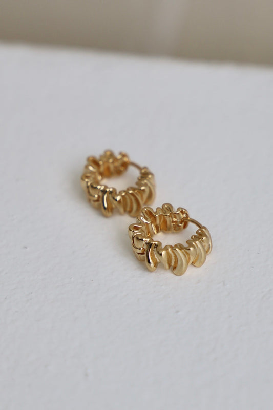 Gold hoop earrings are displayed on a textured white surface. The earrings have a wavy design and are positioned close together, showcasing their shiny finish and intricate details.