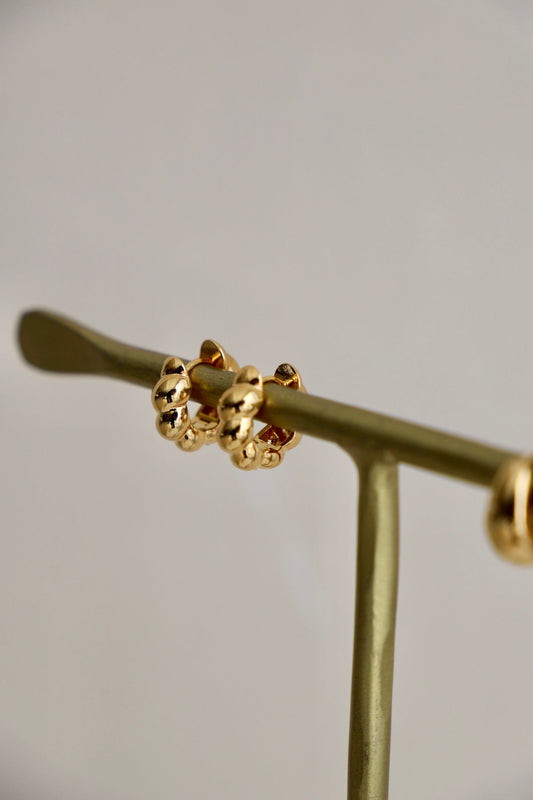 Gold hoop earrings are displayed on a slender, green stand. The earrings rest neatly, showcasing their shiny surface against a soft, neutral background.