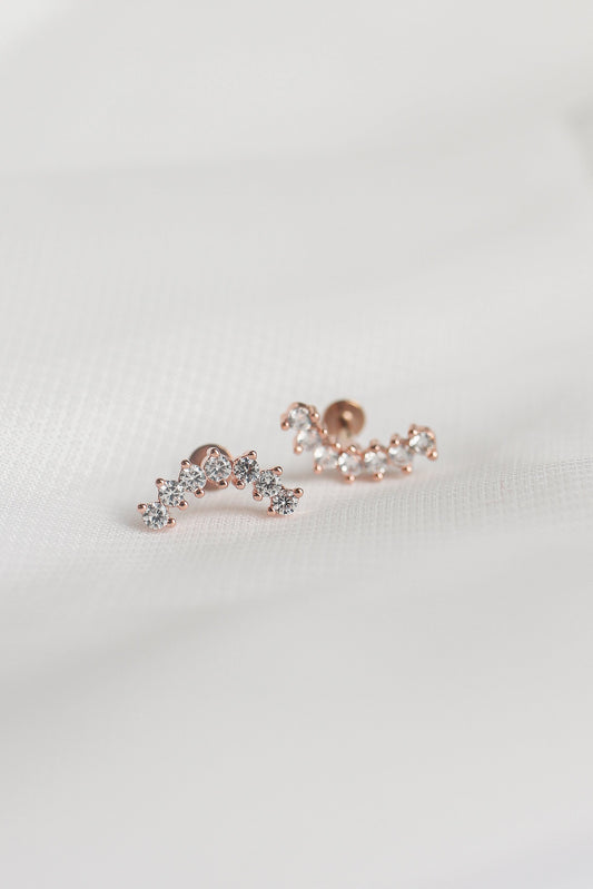 Two rose gold earrings adorned with small, sparkling gemstones are placed on a smooth, white fabric surface, highlighting their elegant design and shine.