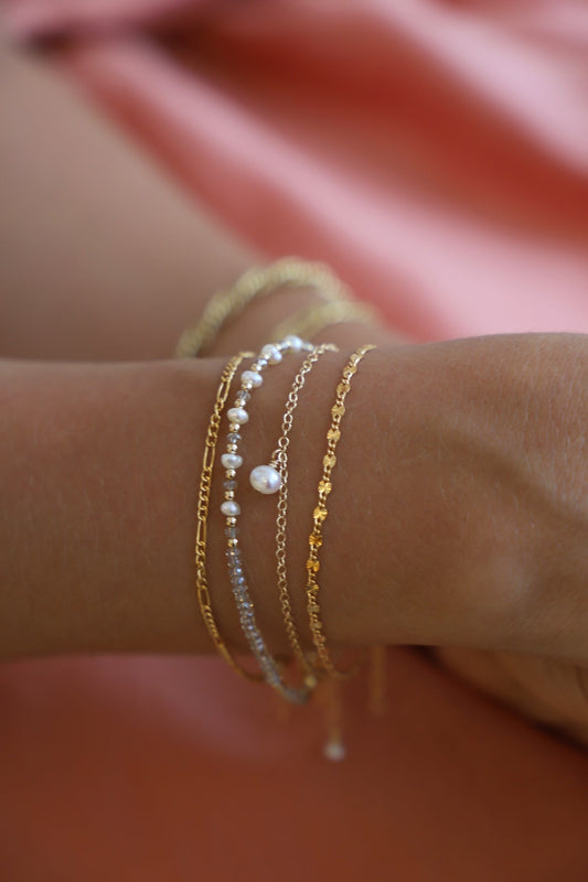 Multiple gold and pearl bracelets adorn a wrist, showcasing delicate designs and varying textures. The background features a soft, pink fabric, enhancing the elegant, feminine aesthetic.