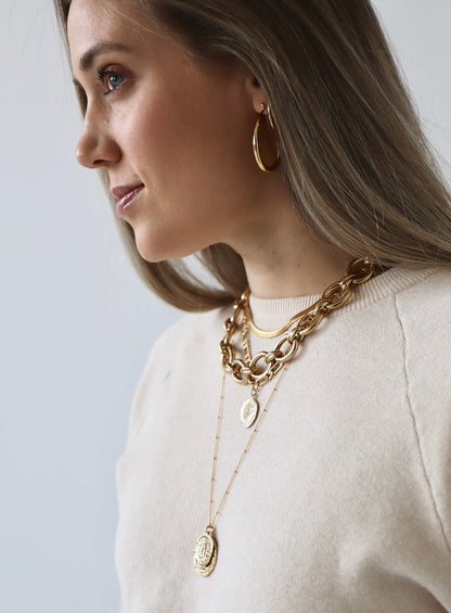 A young woman with long hair wears multiple pieces of gold jewelry. She smiles subtly, posing against a light background, showcasing layered necklaces and hoop earrings.