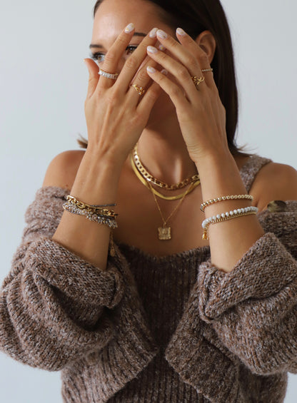 A person displays multiple rings and bracelets while partially covering their face with their hands. They wear a cozy, textured sweater, set against a soft, neutral-colored background.