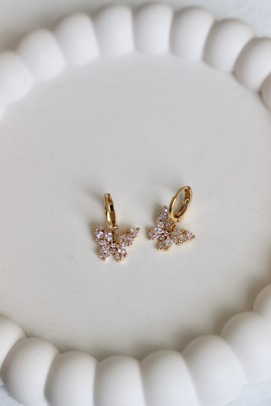 Two butterfly-shaped earrings, adorned with sparkling gemstones, rest on a smooth, white surface. The earrings are highlighted against a softly rounded, pale backdrop.