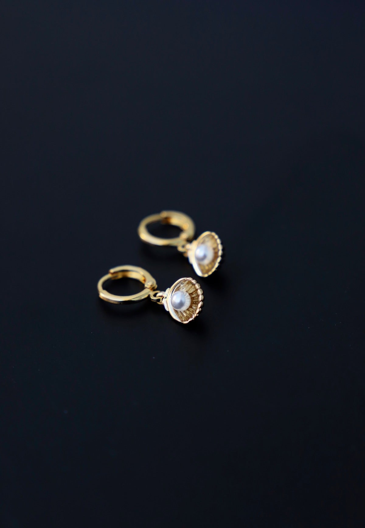 Gold hoop earrings with a shell-like design are displayed, featuring small pearls at the center, resting on a black surface that contrasts with their shiny appearance.