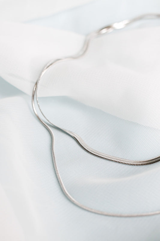 A silver chain rests curved on soft, layered fabric in shades of white and pale blue. The delicate texture of the fabric contrasts with the smooth, shiny surface of the chain.