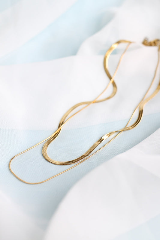 A gold necklace with multiple smooth, curved chains lies atop a soft, pastel blue and white fabric, showcasing its elegance in a serene, well-lit setting.