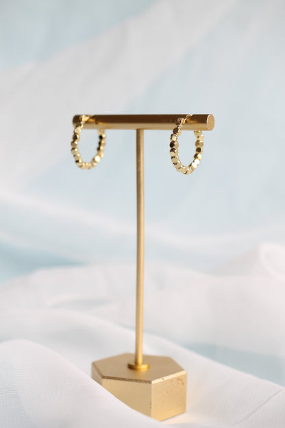 Gold hoop earrings are displayed on a sleek metal stand. The background features soft, light blue and white fabric, creating an elegant and minimalist setting.