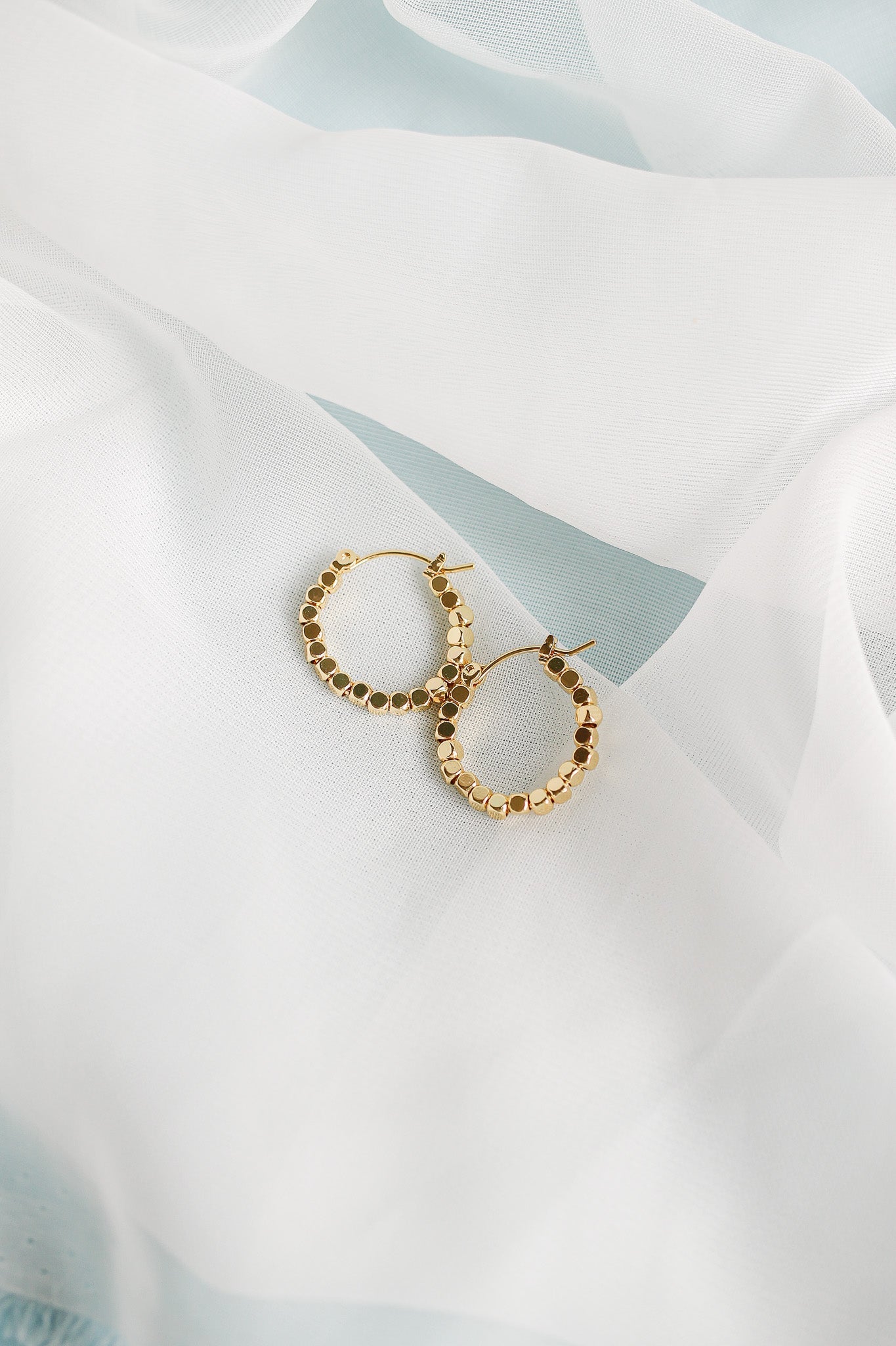 Two golden hoop earrings are delicately resting on a soft, sheer white fabric. The earrings feature a pattern of circular elements, creating an elegant, shimmering effect in a light setting.