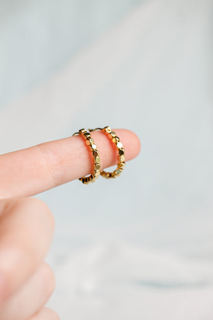 Two gold hoop earrings are held on the tip of a finger, showcasing their shiny, textured surface against a softly blurred light background.