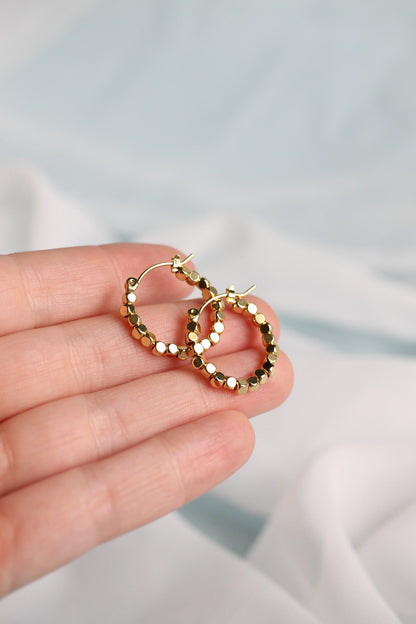 A hand holds a pair of gold hoop earrings featuring small circular embellishments, set against a softly blurred, light-colored background of fabric.