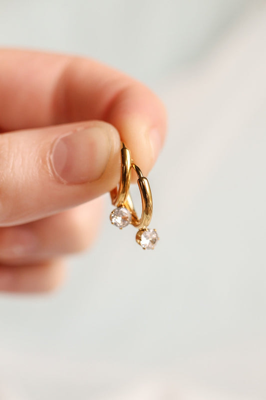 A hand holds a pair of small gold hoop earrings adorned with sparkling clear gemstones, showcasing the jewelry against a softly blurred light background.