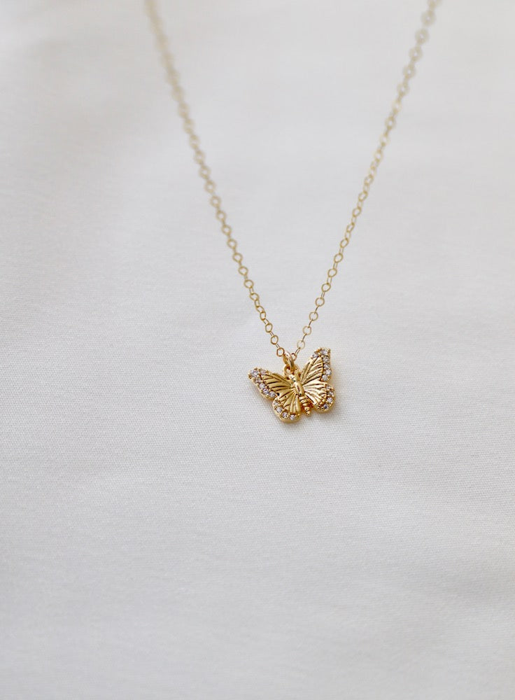 A delicate gold necklace features a butterfly pendant, which hangs gracefully from a fine chain. The necklace rests on a smooth, light-colored fabric background, enhancing its elegance.