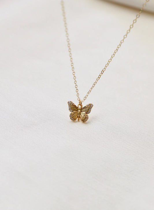 A delicate gold butterfly pendant hangs from a fine chain, resting on a light-colored fabric background, conveying elegance and charm in a softly lit setting.