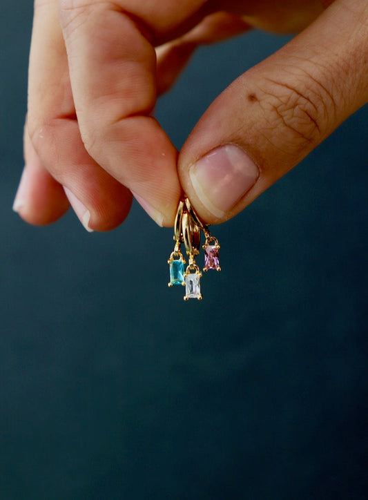 A hand holds three gold hoop earrings, each adorned with different colored gemstonesâ€”blue, clear, and pinkâ€”against a dark, softly blurred background, emphasizing the jewelry's intricate details.