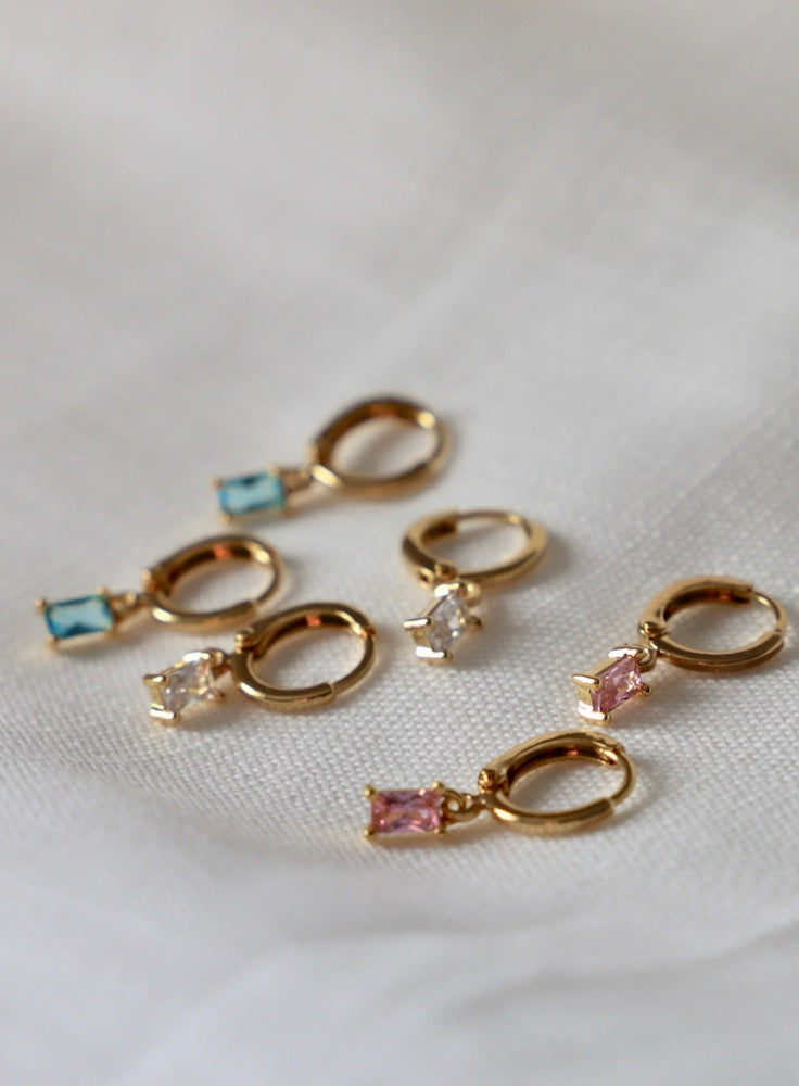 Gold hoop earrings with colored gemstones (blue, clear, pink) rest on a soft, light-colored fabric, showcasing a delicate and elegant arrangement of jewelry pieces.