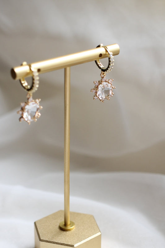 Dangling earrings are showcased on a gold stand. The earrings feature a clear gem surrounded by smaller pink stones, set against a soft, blurred fabric backdrop.