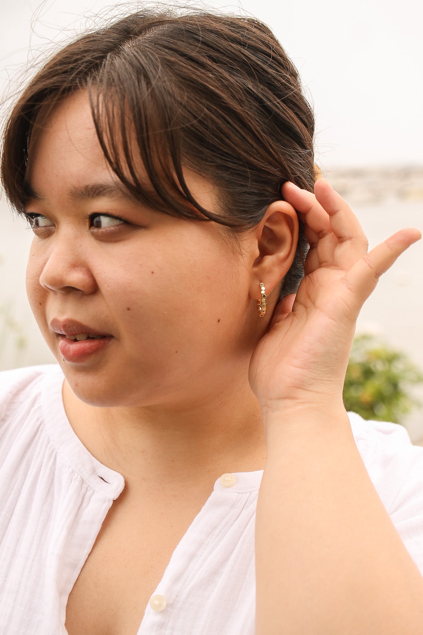 A woman with shoulder-length hair is touching her ear while looking thoughtfully to the side. She wears a white blouse and appears outdoors, with a blurred natural background.