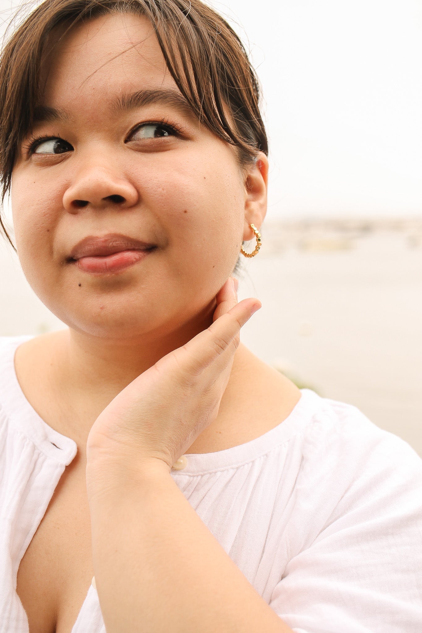 A young woman with short hair wears gold hoop earrings, gently touching her neck while looking to her side, against a soft, neutral background suggesting an outdoor setting.