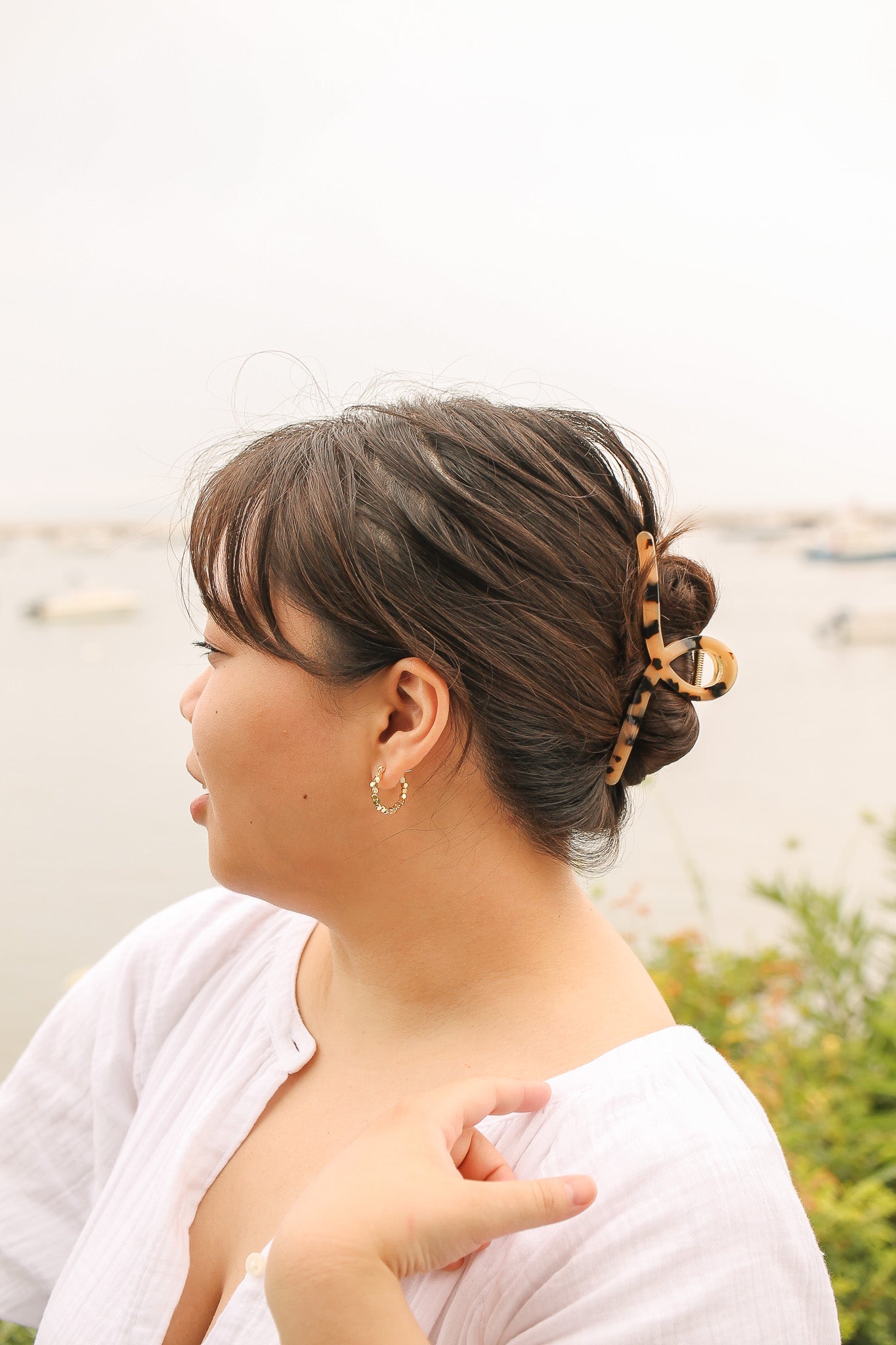 A woman with a bun hairstyle, secured by a patterned clip, is looking away towards a calm body of water. She has a hand on her shoulder, surrounded by greenery.